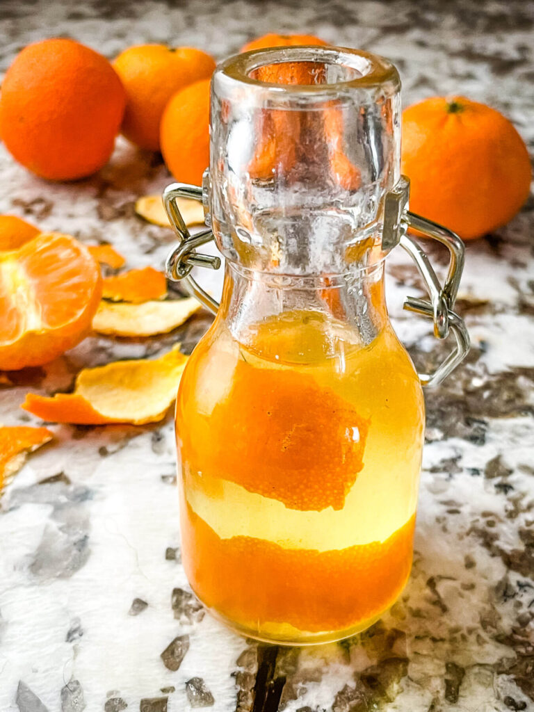 A small bottle full of Orange Simple Syrup with peels, and oranges behind it