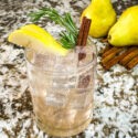 A clear glass with the Pear Tree Vodka Cocktail and garnished with pear, rosemary, and cinnamon stick