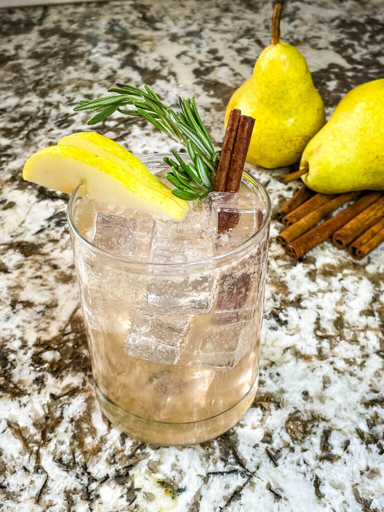 A clear glass with the Pear Tree Vodka Cocktail and garnished with pear, rosemary, and cinnamon stick