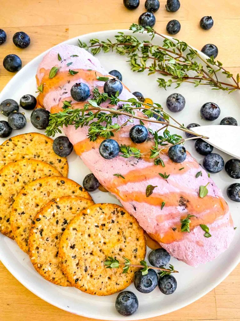 The log of Blueberry Goat Cheese on a white plate with crackers, berries, and thyme around it