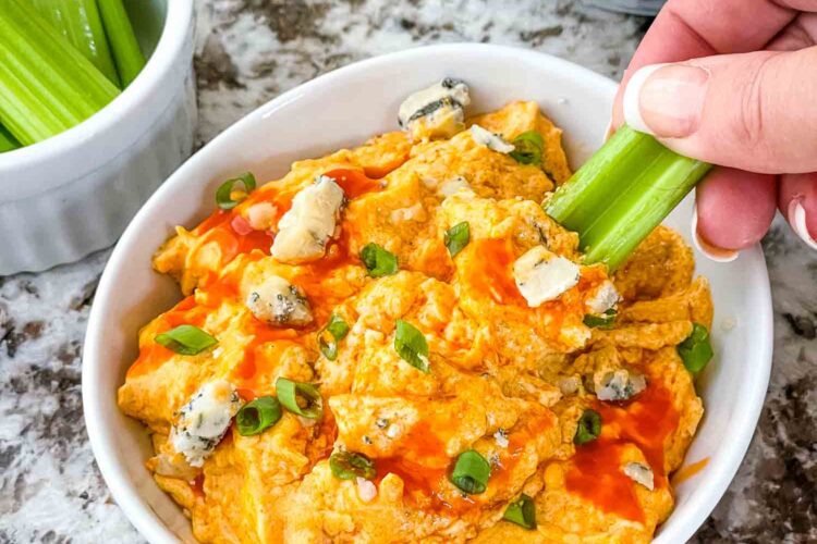 A hand scooping celery into a bowl of Buffalo Chicken Crockpot Dip