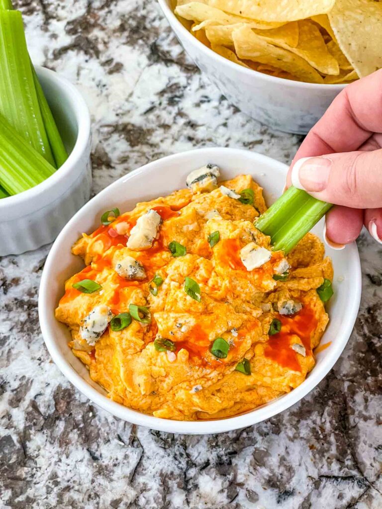 A hand scooping celery into a bowl of Buffalo Chicken Crockpot Dip