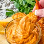 A hand dunking a breaded shrimp into a small bowl of Cajun Sauce