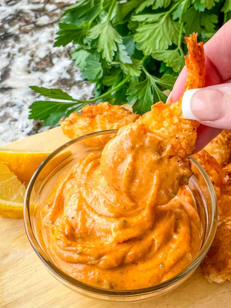 A hand dunking a breaded shrimp into a small bowl of Cajun Sauce