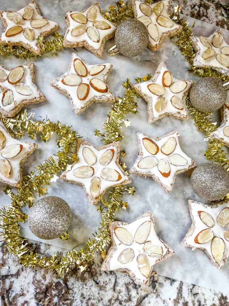 Zimmerstern (German Cinnamon Star Cookies) on parchment paper with a few silver ornament and a gold garland woven throughout