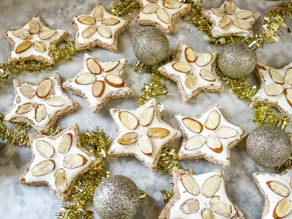 Zimmerstern (German Cinnamon Star Cookies) on parchment paper with a few silver ornament and a gold garland woven throughout