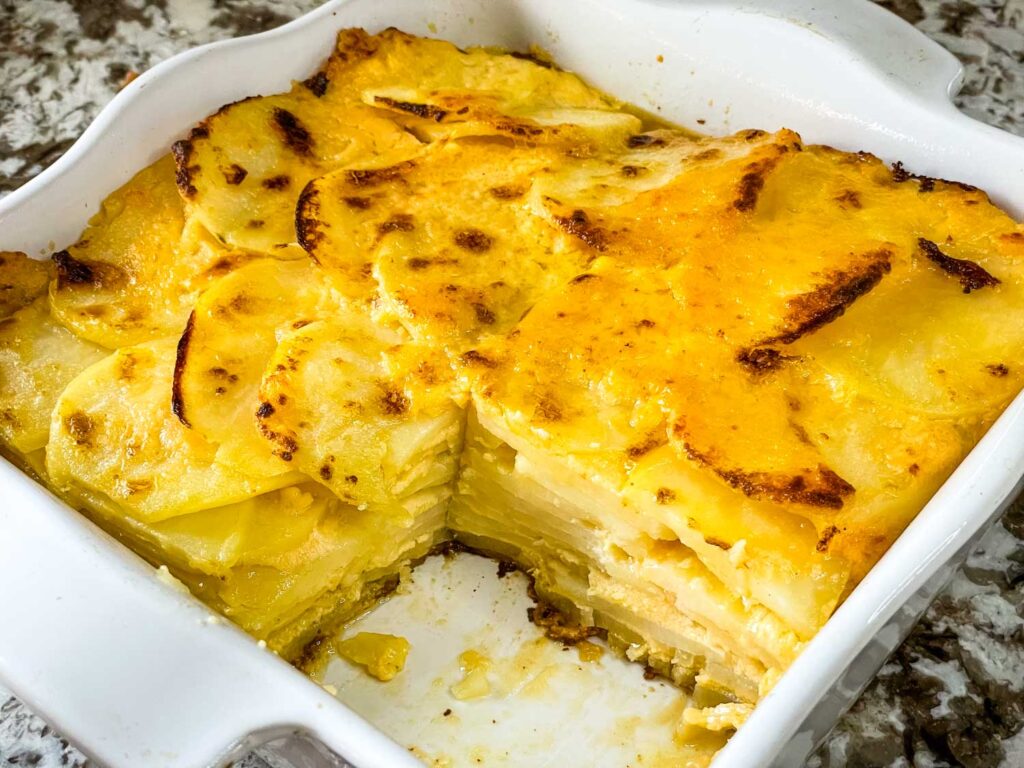 A square white dish full of Scalloped Cheesy Potatoes with a corner cut out of it