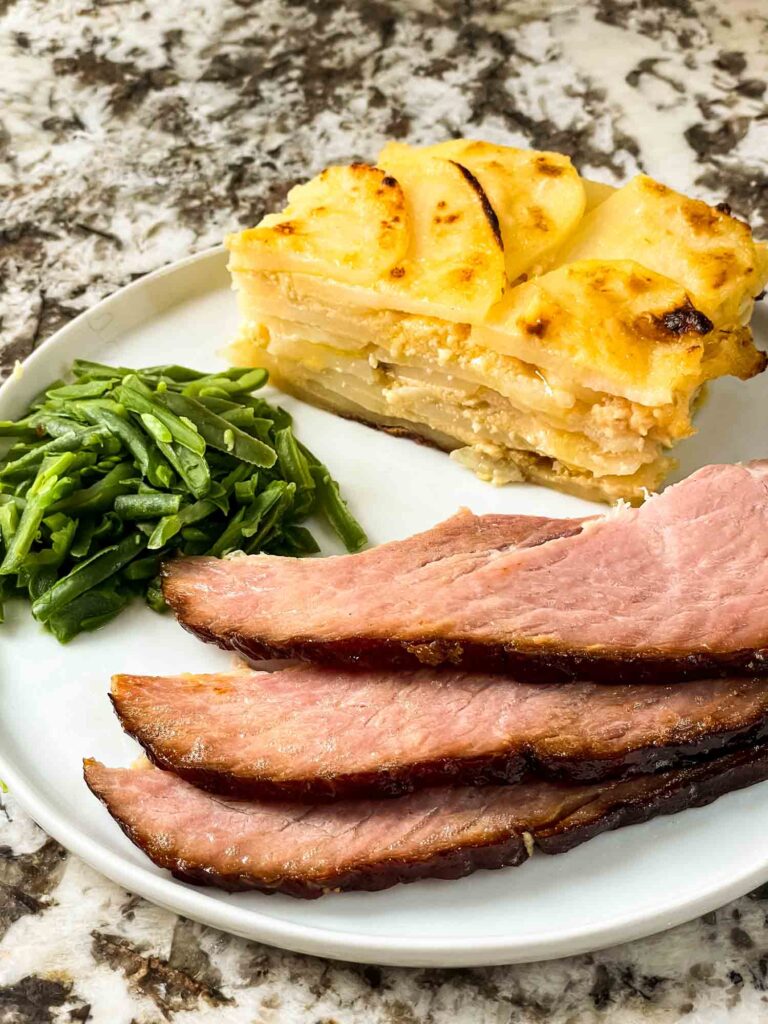 Slices of Spiral Ham with Brown Sugar Glaze on a plate with some green beans and cheesy scalloped potatoes