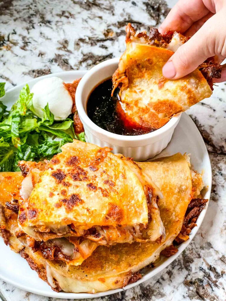 A hand dunking a Birria Quesadilla into au jus