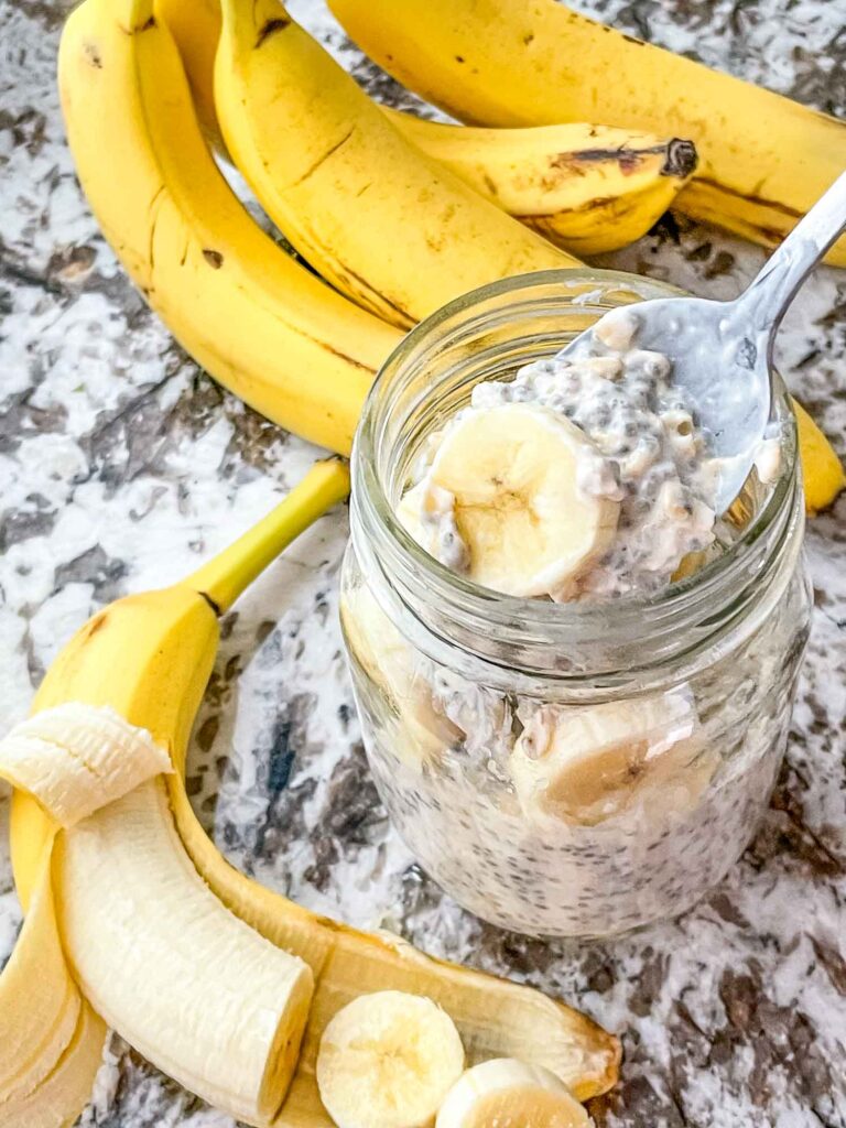 A spoon digging into a mason jar of Banana Overnight Oats with banana around it