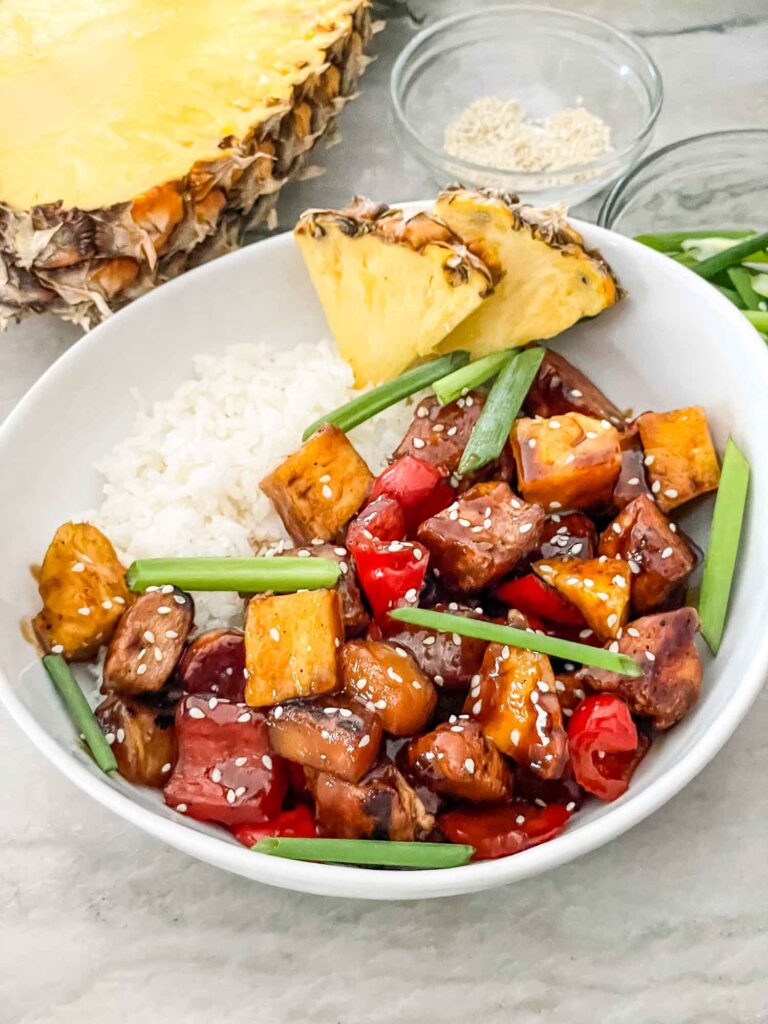 A white bowl filled with white rice and Pineapple Chicken surrounded by pineapple pieces