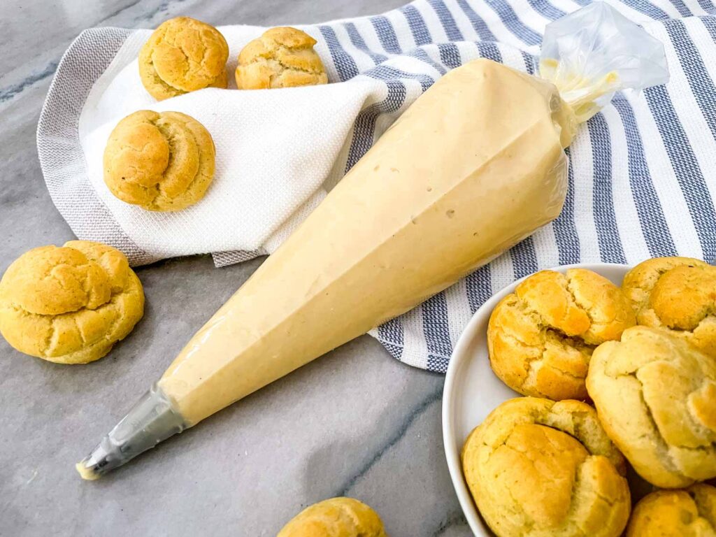 Vanilla Pastry Cream in a piping bag surrounded by cream puffs and on a towel