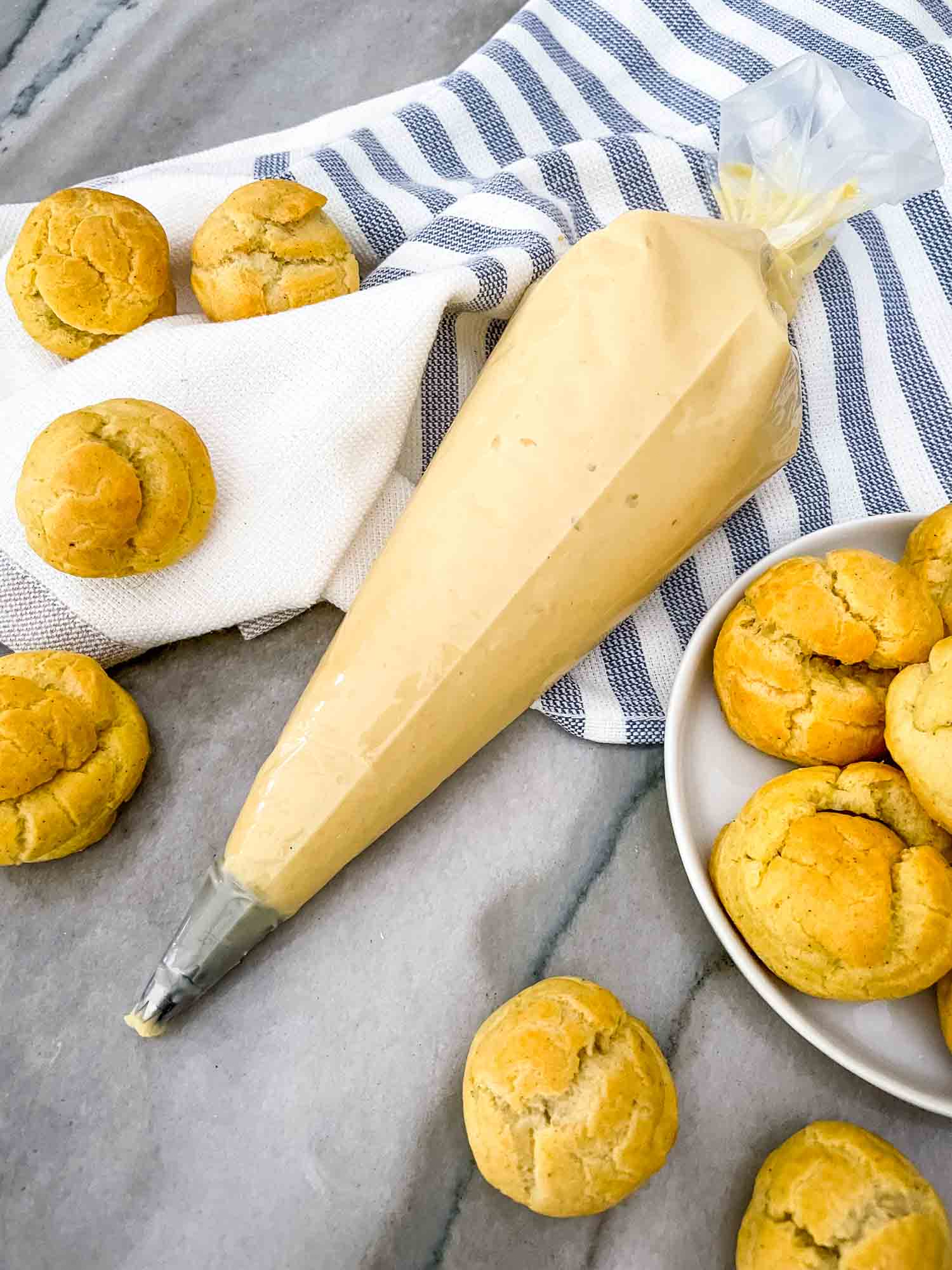 A piping bag full of Vanilla Pastry Cream and surrounded by cream puffs on a blue and white towel