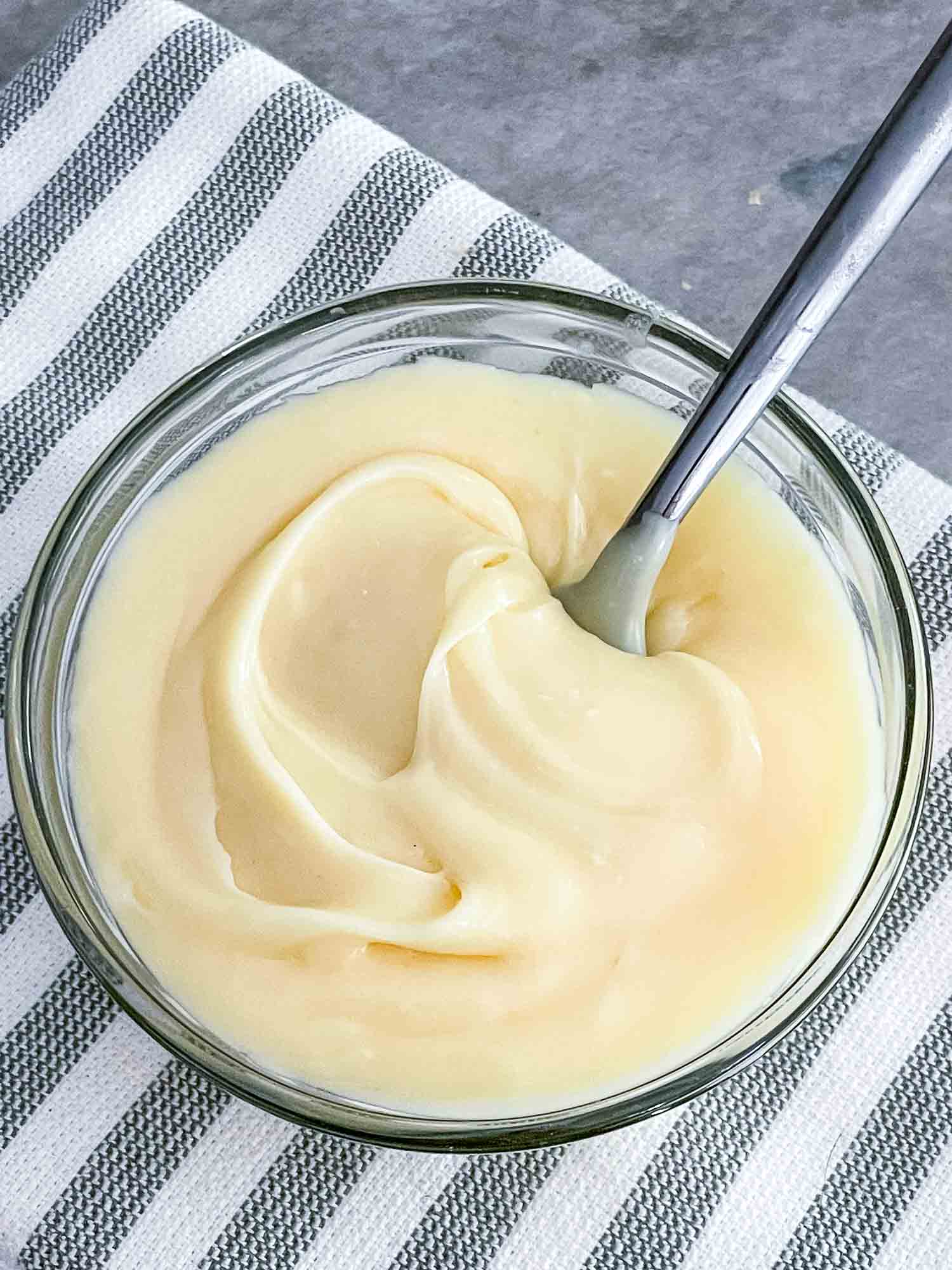 Top view of White Chocolate Ganache in a bowl with a spoon in it, sitting on a striped towel