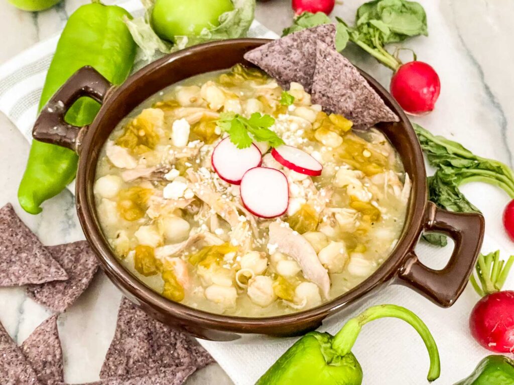 Chicken and Hatch Green Chile Pozole in a brown bowl surrounded by toppings