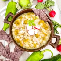 Chicken and Hatch Green Chile Pozole in a brown bowl with topping ingredients around it
