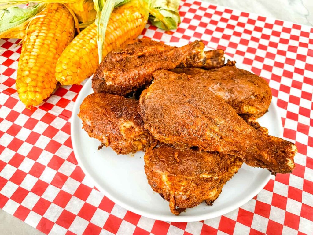 A plate of Smoked Chicken Legs next to smoked corn on a red checkered mat