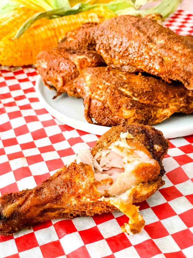 A partially eaten leg with a plate of Smoked Chicken Legs behind it sitting on a red checkered paper