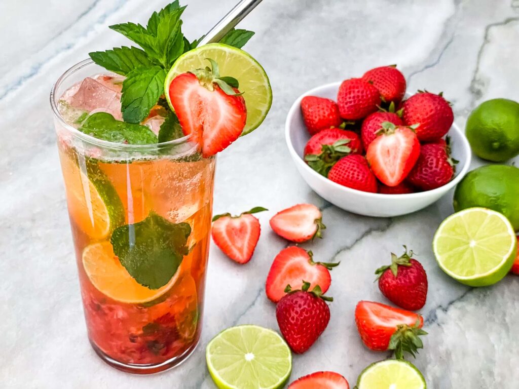 A tall glass of Strawberry Mojito with limes and strawberries next to it