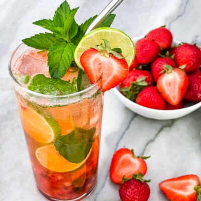 A tall glass of Strawberry Mojito with berries and limes around it and a silver straw