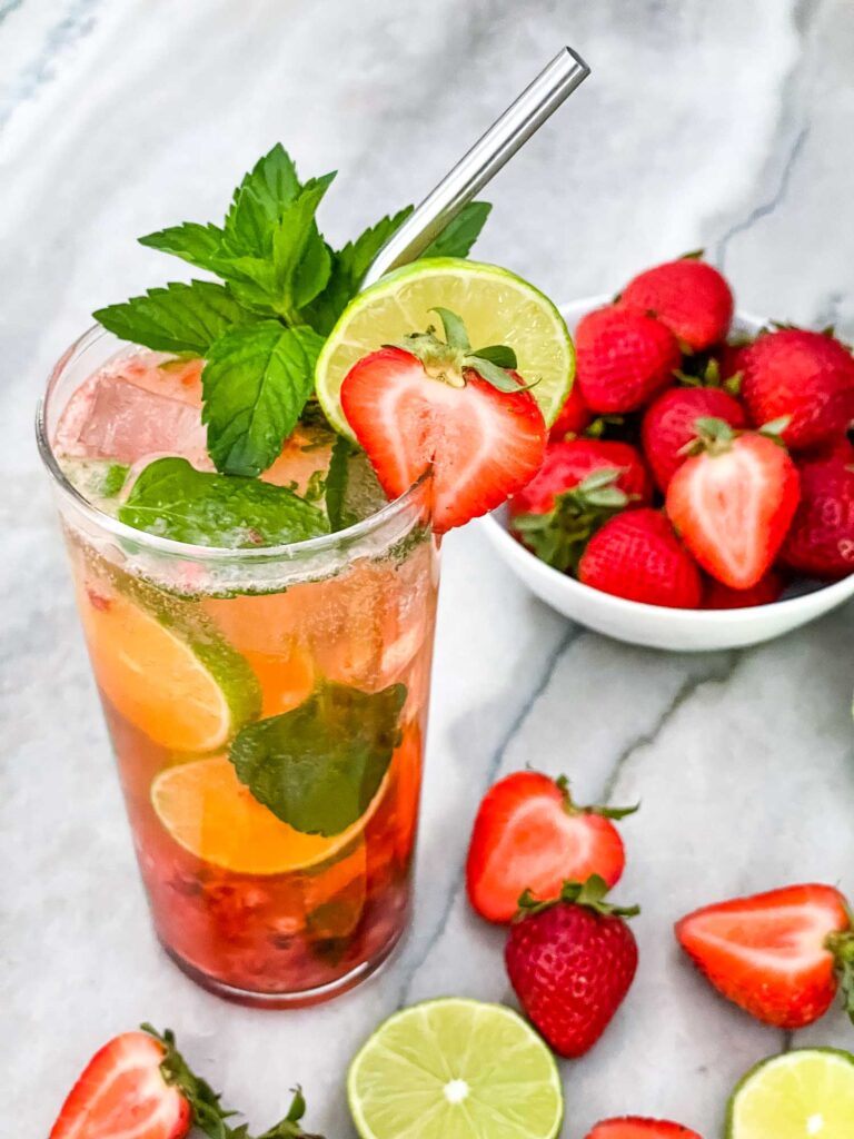 A tall glass of Strawberry Mojito with berries and limes around it and a silver straw