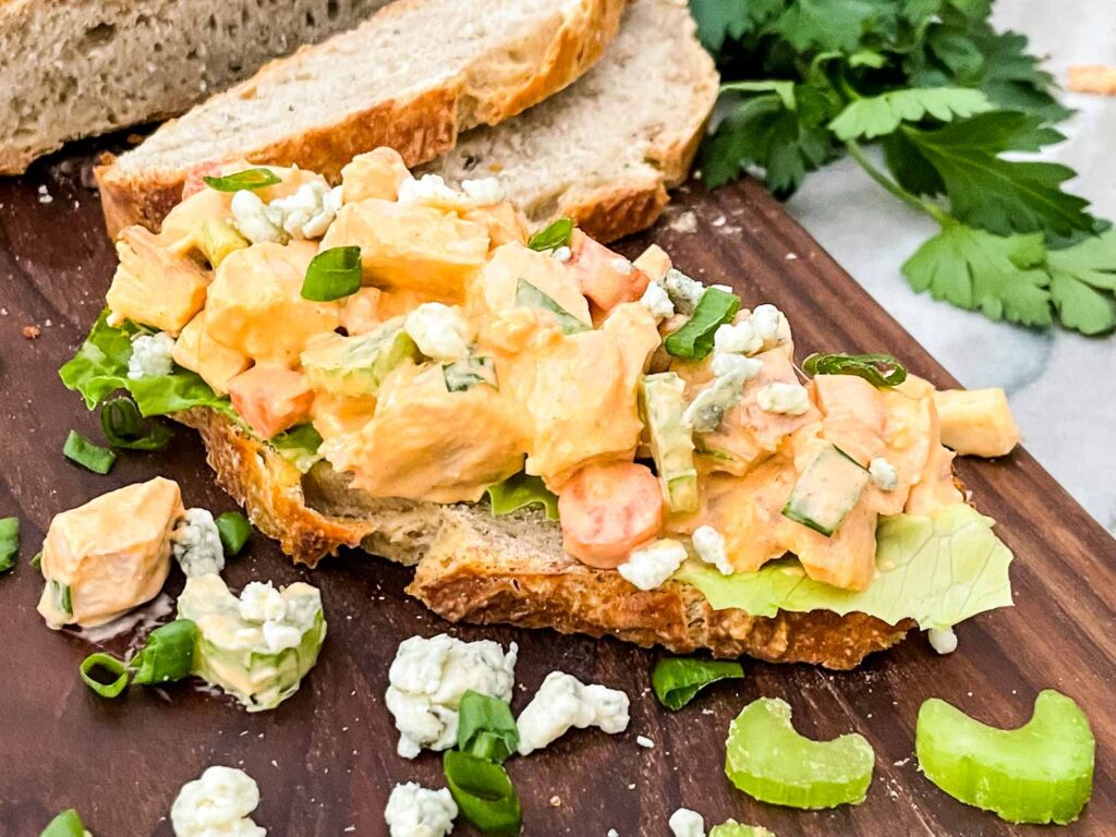 A piece of bread topped with Buffalo Chicken Salad and sitting on a dark piece of wood