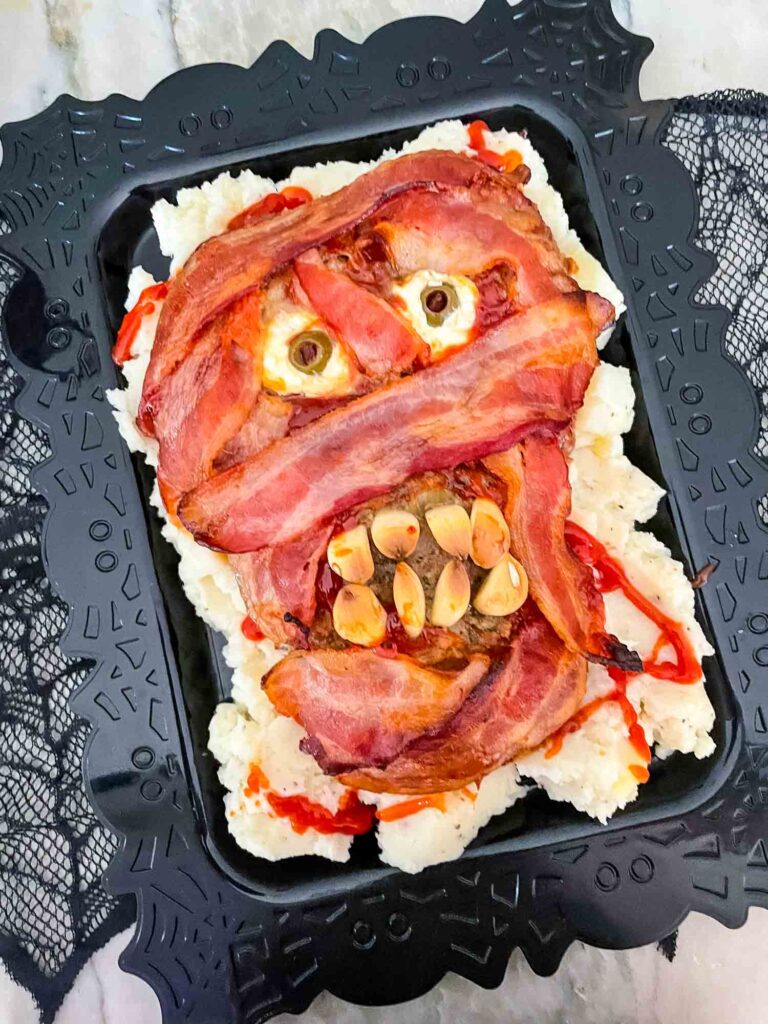 The Halloween Mummy Meatloaf on a black platter with mashed potatoes under it