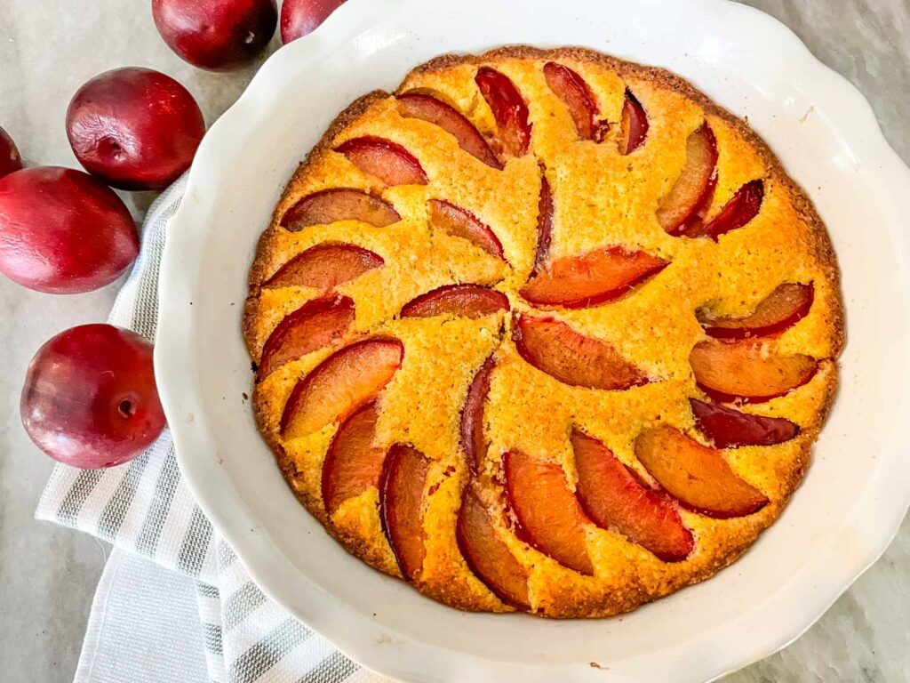 A Plum Cake in a white pie dish with plums next to it