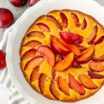 Top view of a Plum Cake in a white pie dish on a towel with whole plums next to it