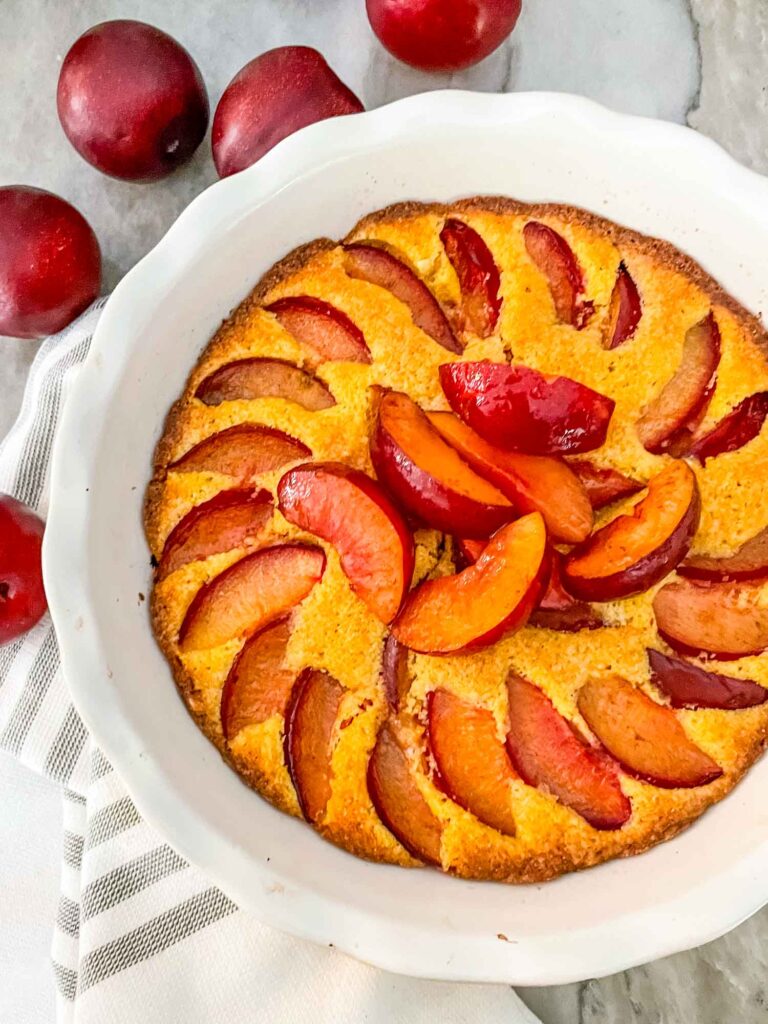 Top view of a Plum Cake in a white pie dish on a towel with whole plums next to it