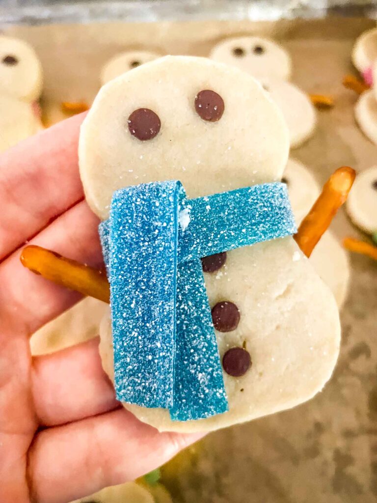 A hand holding a Shortbread Snowman Cookie with a blue scarf