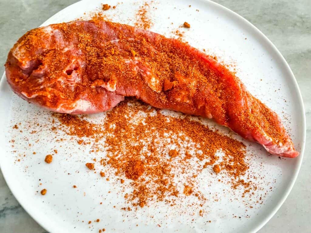The meat on a white plate with the rub being applied