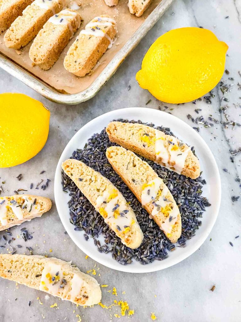 Lavender Lemon Biscotti on top of lavender buds on a small plate with more cookies and lemons around it