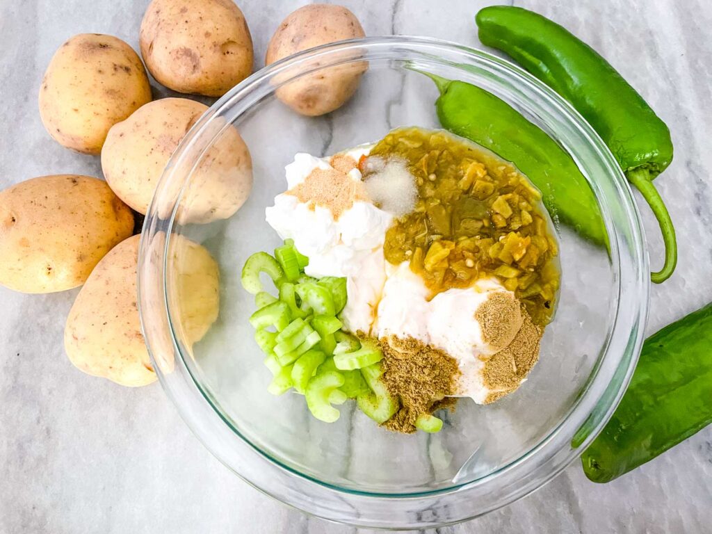 The sauce ingredients in a clear glass bowl with fresh peppers and whole potatoes on the counter next to it