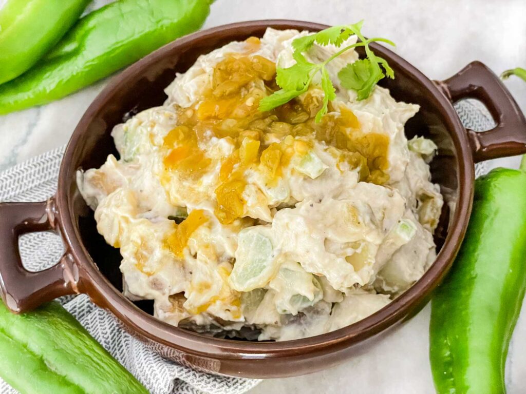 Hatch Green Chile Potato Salad in a brown shallow bowl with small handles. Sitting on a counter with fresh green peppers around it