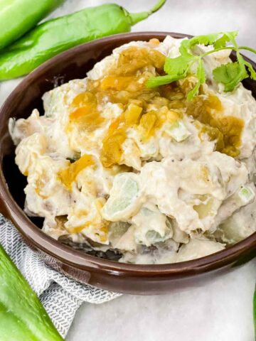 Hatch Green Chile Potato Salad piled up in a brown bowl with diced peppers on top. Sitting on a counter with fresh green peppers around it