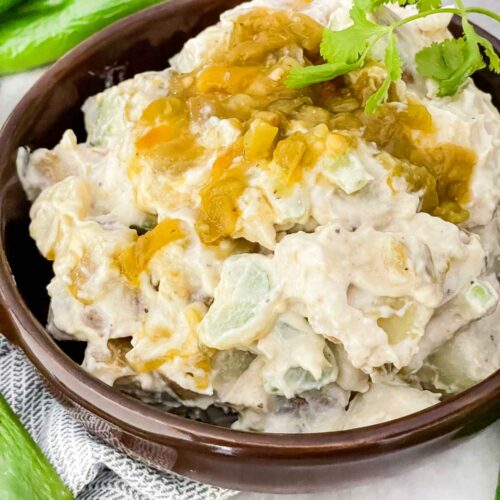 Hatch Green Chile Potato Salad piled up in a brown bowl with diced peppers on top. Sitting on a counter with fresh green peppers around it