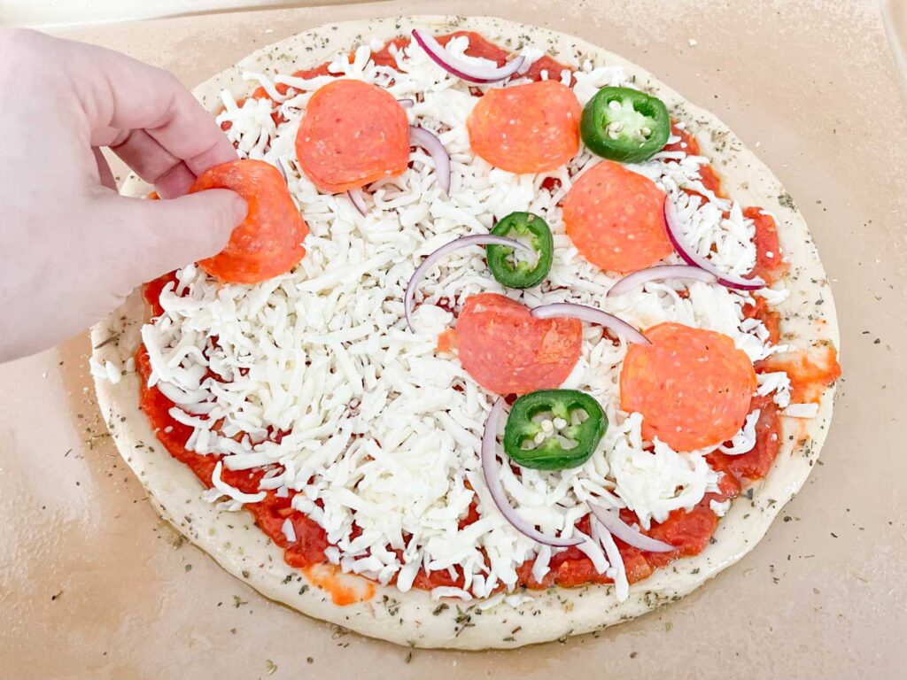 A hand adding toppings on top of a pizza with sauce and cheese before baking