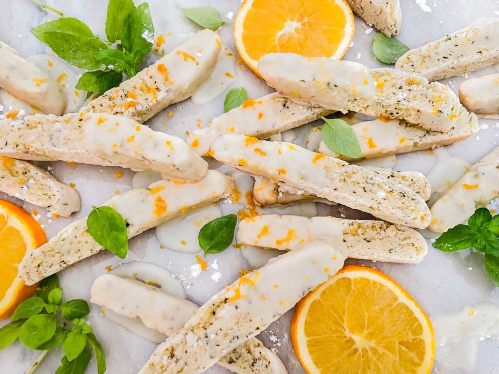 A pile of Basil Orange Biscotti cookies on a counter with fresh basil and oranges