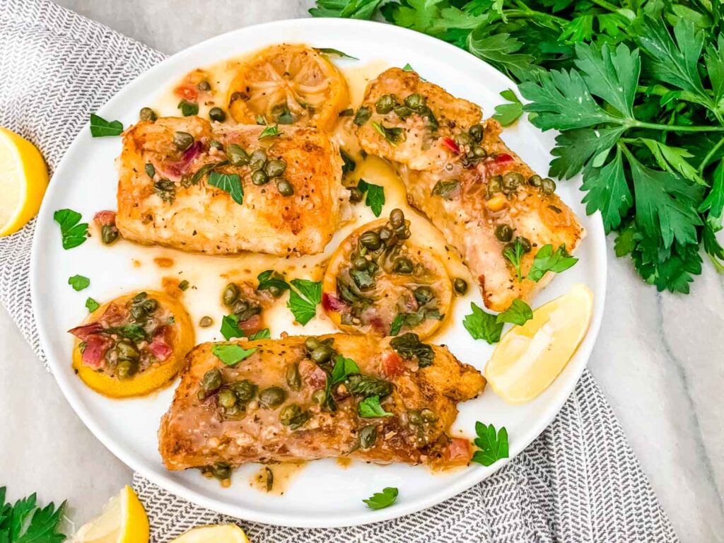 Three pieces of Cod Piccata on a white round plate with lemon and parsley garnishes