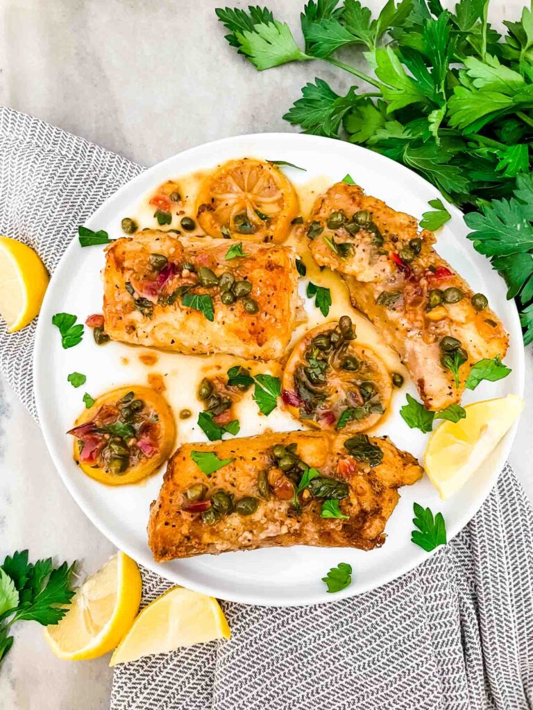 Top view of a large white plate with three pieces of Cod Piccata. Lemon and parsley around the plate.