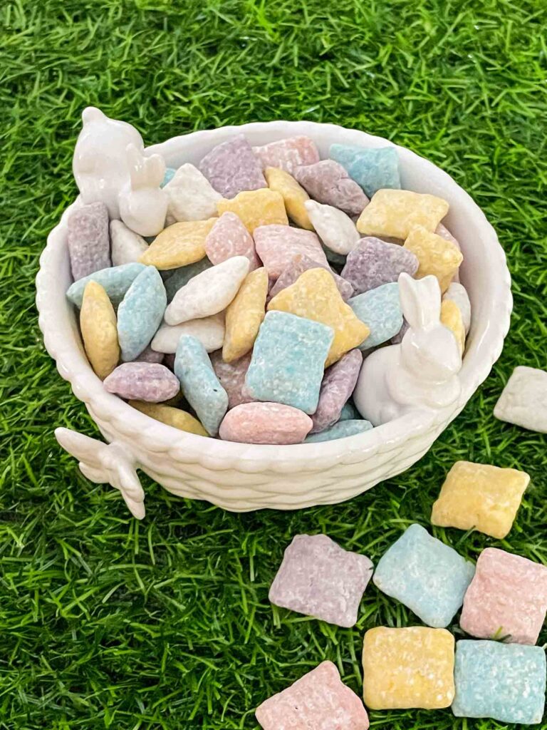 A small white bowl with two bunnies full of Easter Puppy Chow sitting on green grass