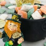 Pin of Leprechaun Puppy Chow in a small black cauldron in white, green, and orange. With a leprechaun standing next to it.