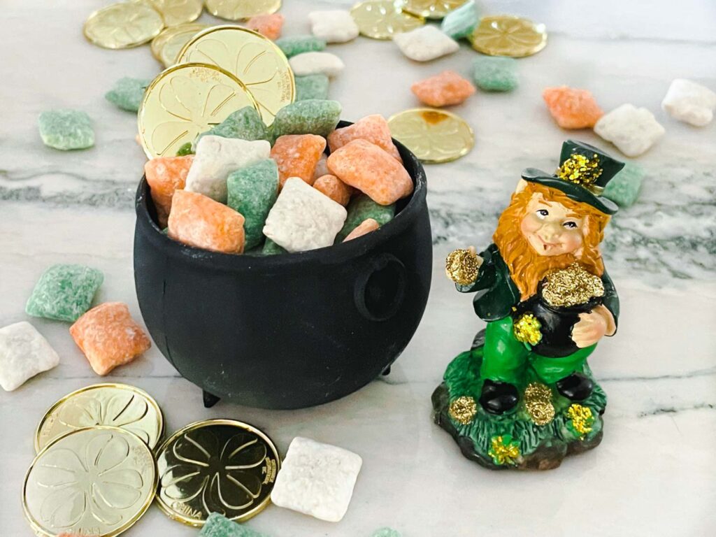 A small black cauldron full of Leprechaun Puppy Chow next to a leprechaun with more muddy buddies and gold coins scattered around them