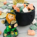 A leprechaun standing in front of a small cauldron full of Leprechaun Puppy Chow with more candy and gold coins around it