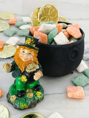 A leprechaun standing in front of a small cauldron full of Leprechaun Puppy Chow with more candy and gold coins around it