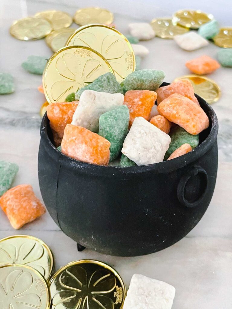 A small black cauldron filled with Leprechaun Puppy Chow in orange, white, and green with more food and gold coins around it