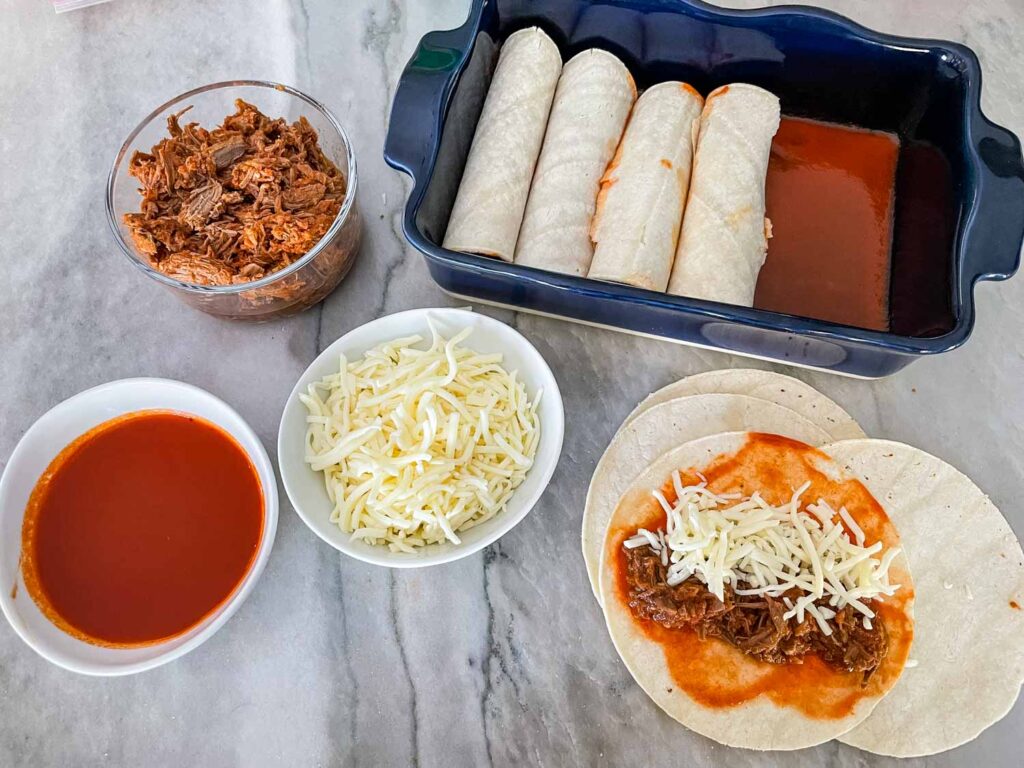 Assembly of the enchiladas on a counter. Tortillas being filled, a bowl of cheese, a bowl of sauce, a bowl of beef, and the baking dish half full of rolled enchiladas