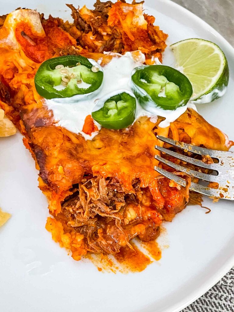 A fork cutting into two Shredded Beef Enchiladas on a white plate with sour cream, lime, and peppers