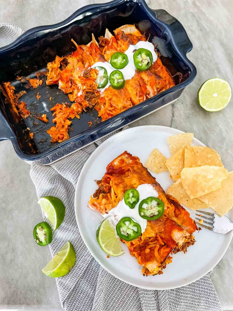 Top view of the finished dish of Shredded Beef Enchiladas with some missing that have been put on a white plate. Surrounded by chips, limes, and peppers.